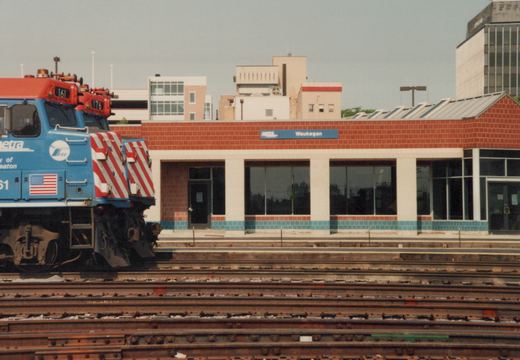 Two blue Metra locomotives with red roofs are on multiple tracks at the Waukegan station, which has a red brick building with a blue sign.