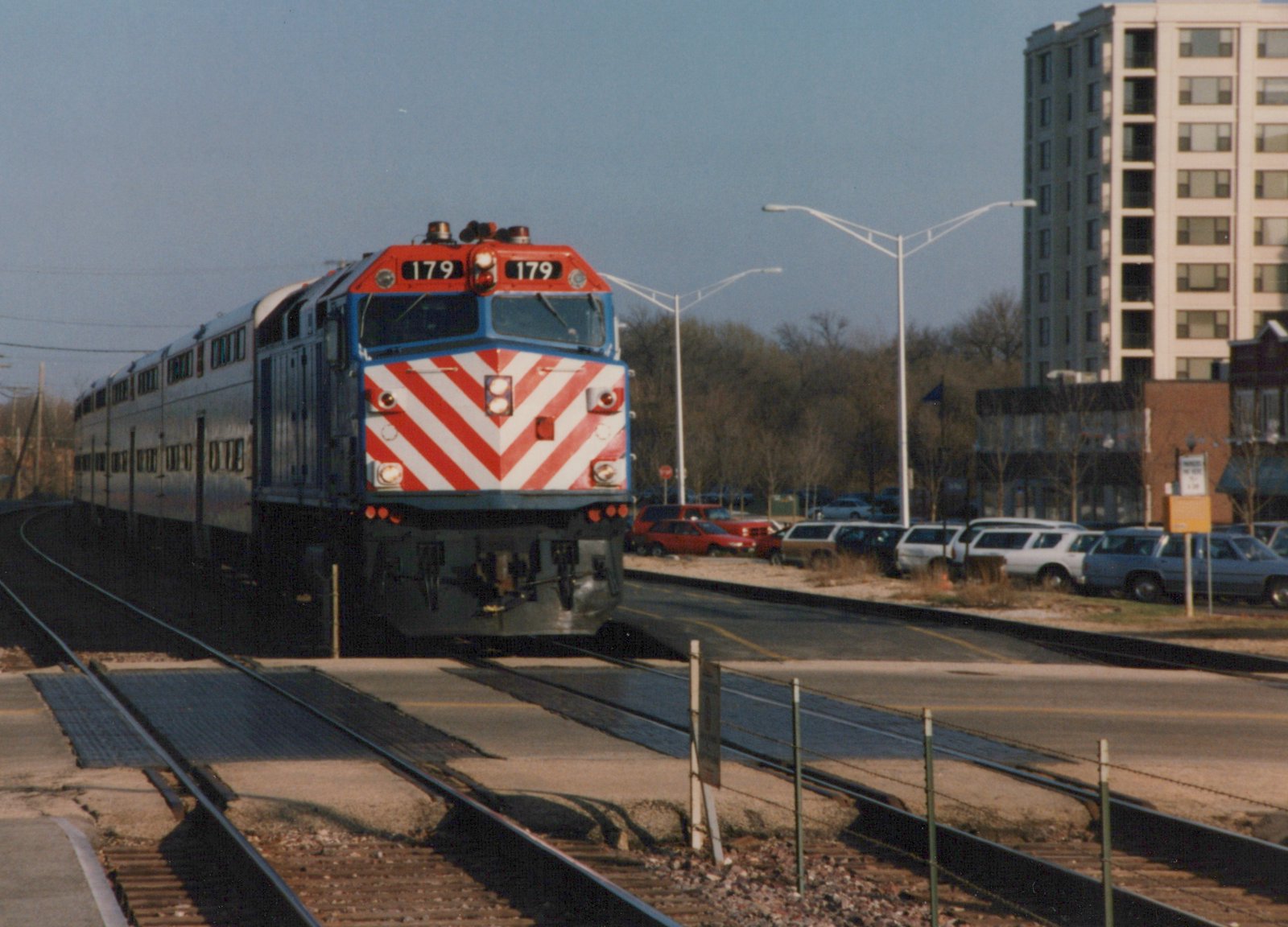 A red, white and blue Metra locomotive numbered 179 pulls silver double-decker commuter cars along tracks that cross a street, with bare trees and parked cars in the background.