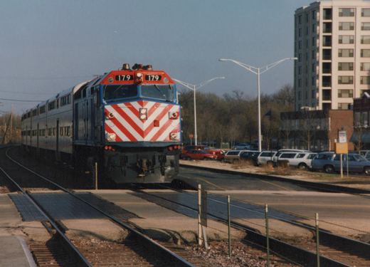 A red, white and blue Metra locomotive numbered 179 pulls silver double-decker commuter cars along tracks that cross a street, with bare trees and parked cars in the background.