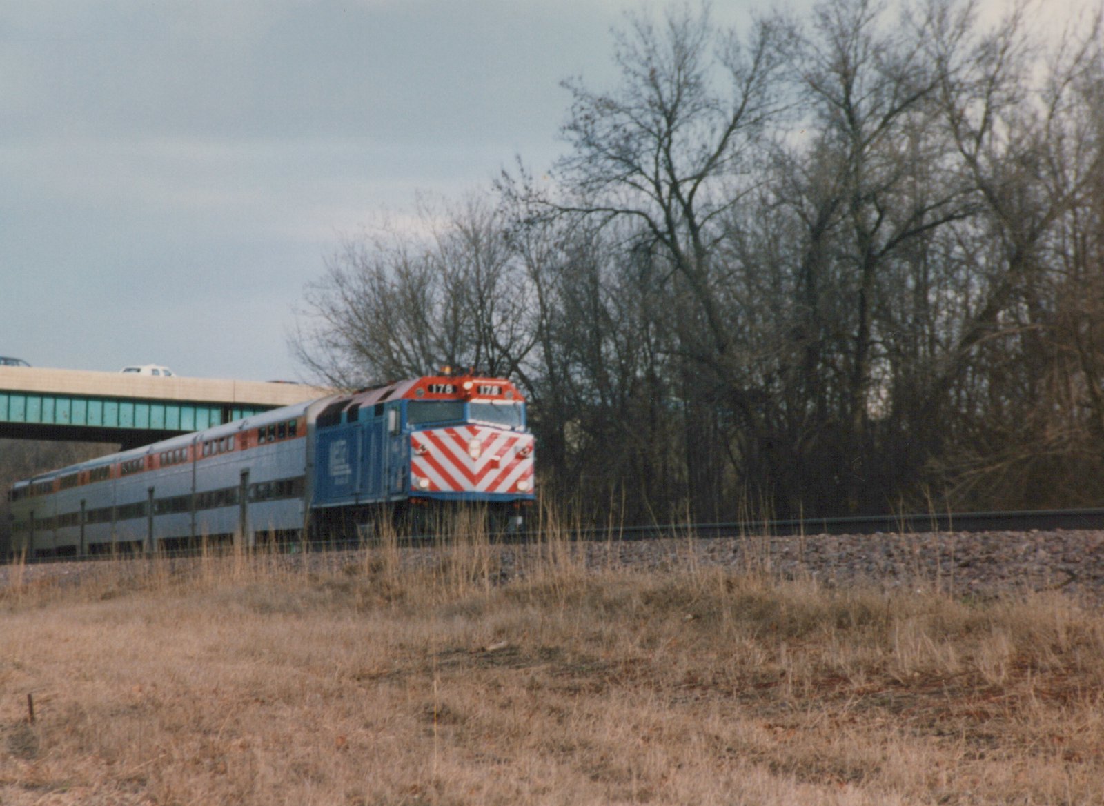 A blue Metra locomotive with a red and white chevron nose, number 178, pulls a bi-level train through a leafless winter landscape with a highway overpass in the background.