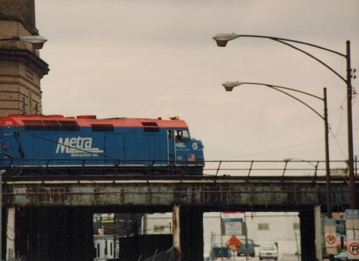 A blue and red Metra locomotive numbered 174 is on an elevated steel bridge in an urban setting under an overcast sky.