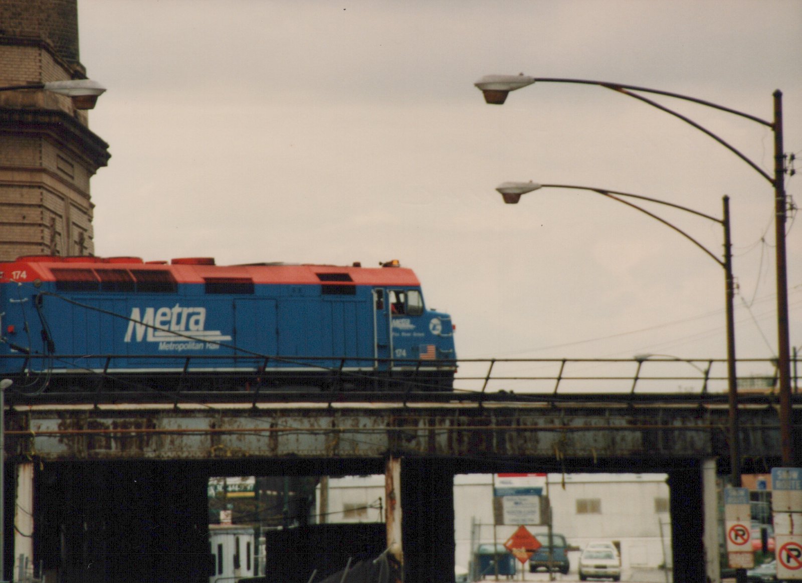 A blue and red Metra locomotive numbered 174 is on an elevated steel bridge in an urban setting under an overcast sky.