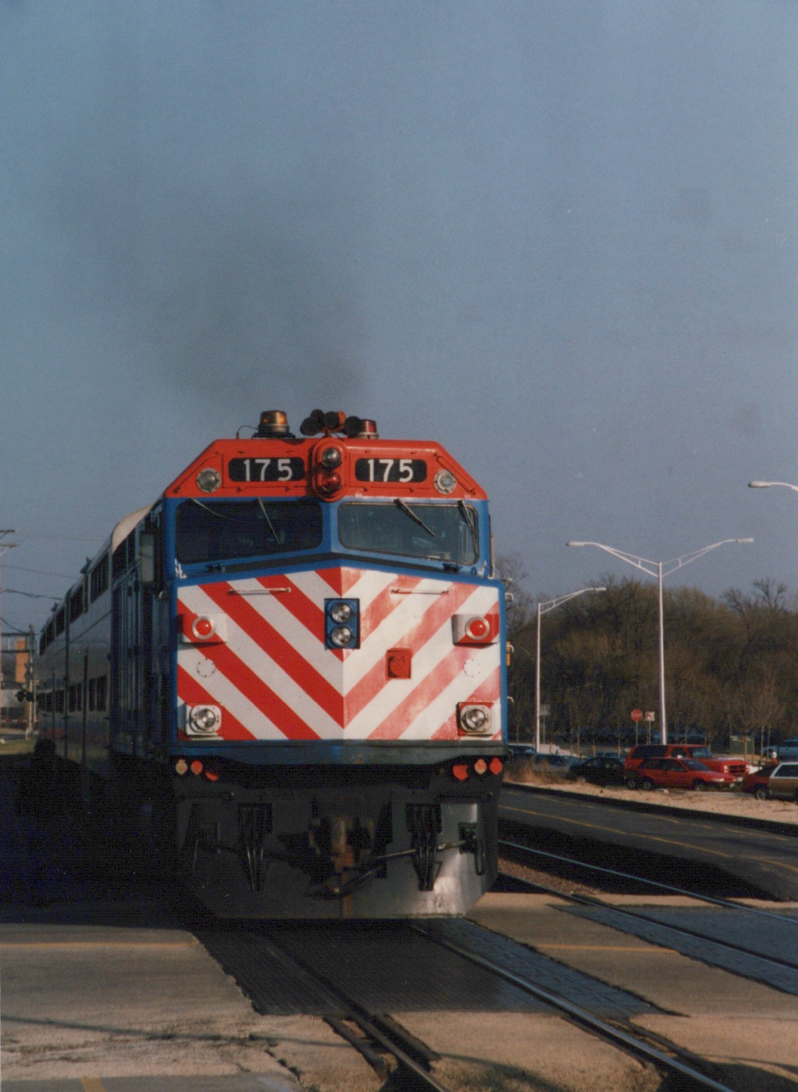 Front view of a red, white, and blue Metra locomotive numbered 175 moving along tracks at a grade crossing, with cars and trees in the background.