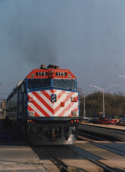 Front view of a red, white, and blue Metra locomotive numbered 175 moving along tracks at a grade crossing, with cars and trees in the background.
