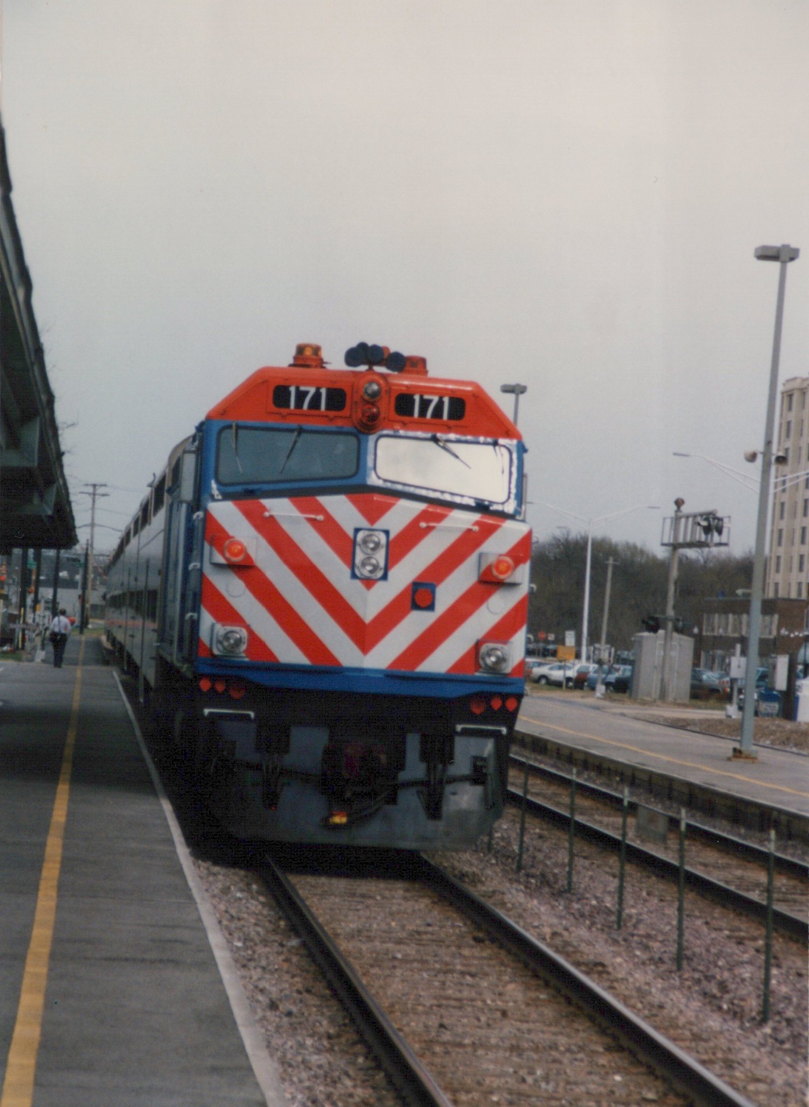 A red, white and blue diesel locomotive numbered 171, identified as a Metra EMD F40PH, is stopped at a station platform with a person walking nearby.