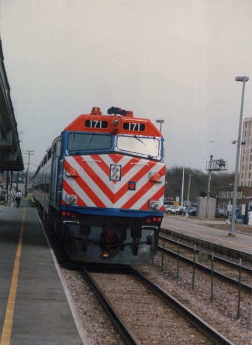 A red, white and blue diesel locomotive numbered 171, identified as a Metra EMD F40PH, is stopped at a station platform with a person walking nearby.