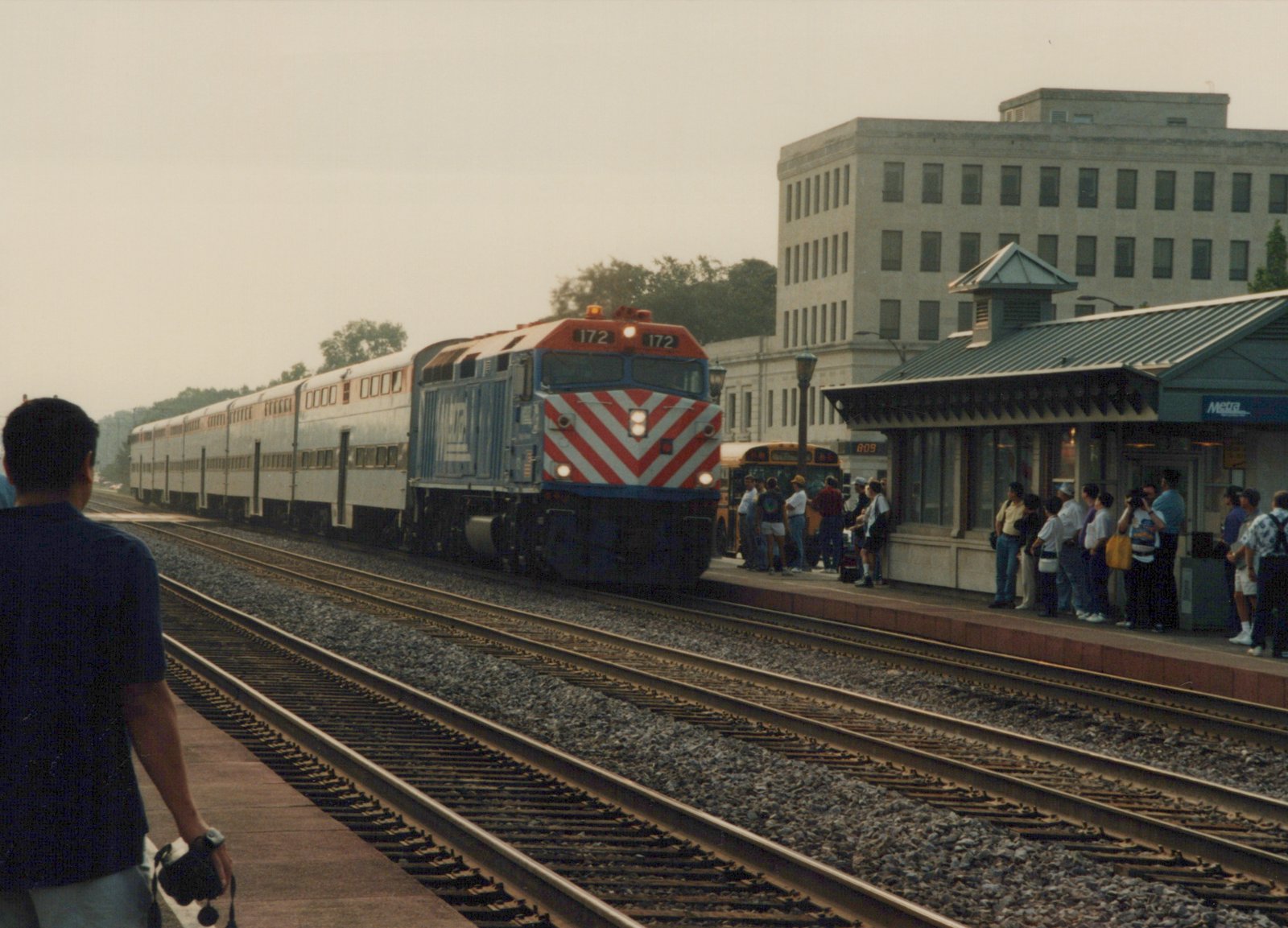 A Metra train with locomotive 172 and silver BiLevel coaches is stopped at a station platform where passengers are waiting, with a school bus visible in the background.