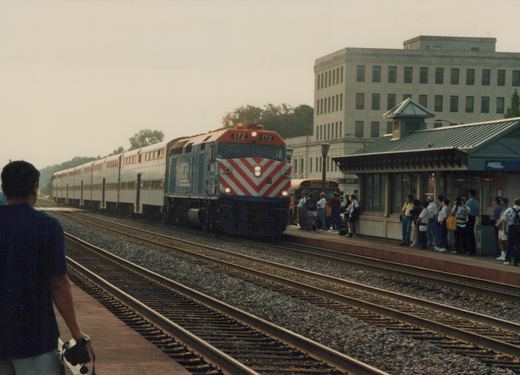 A Metra train with locomotive 172 and silver BiLevel coaches is stopped at a station platform where passengers are waiting, with a school bus visible in the background.