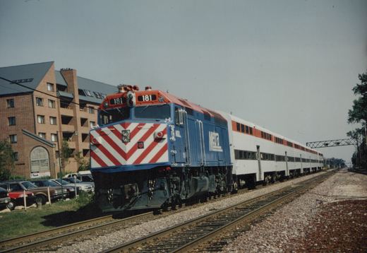 A blue and red Metra diesel locomotive with the number 181 pulls a long train of white double-decker commuter cars on a track next to a brick building and a parking lot.