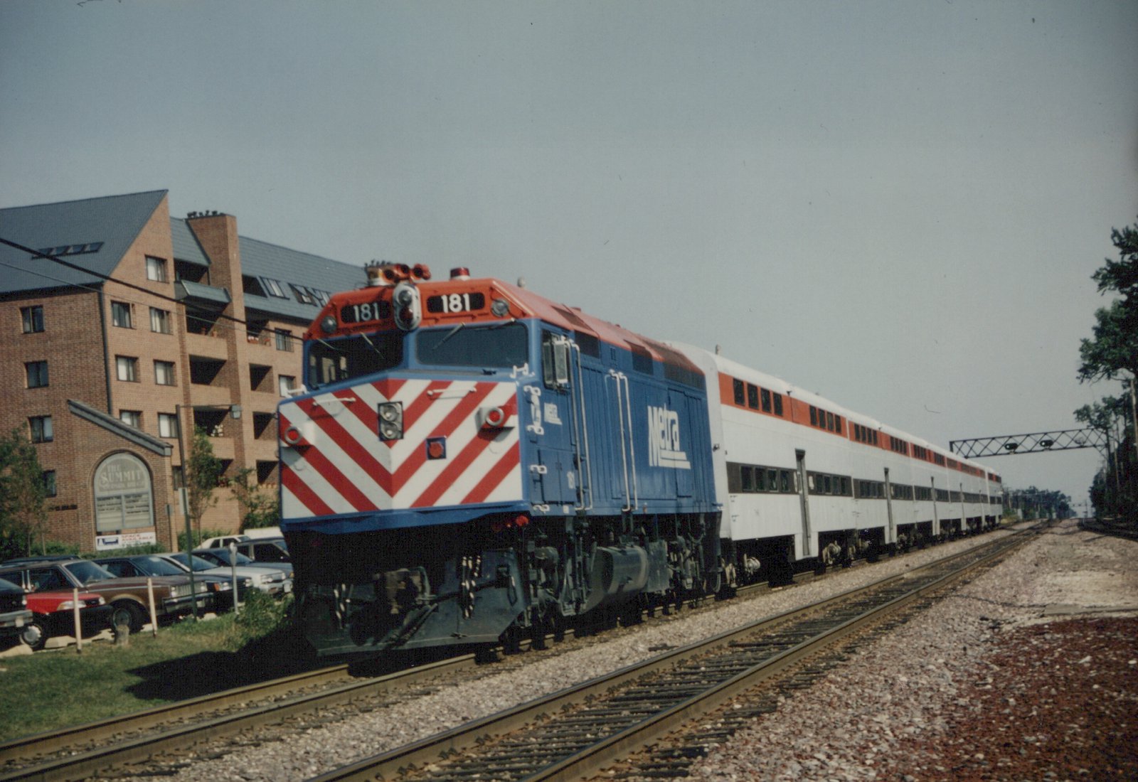 A blue and red Metra diesel locomotive with the number 181 pulls a long train of white double-decker commuter cars on a track next to a brick building and a parking lot.