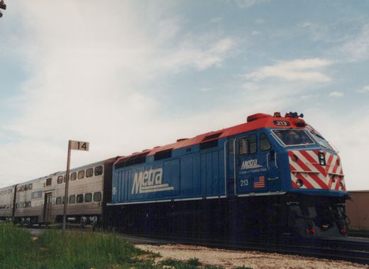 A blue and red Metra locomotive numbered 213 pulls a train of silver passenger cars along a track beside a grassy field under a cloudy sky.