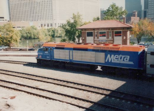 A blue and orange Metra diesel locomotive, number 600, is stopped at the Lake Street station platform, with a small shelter and city skyscrapers visible behind it on a clear autumn day.