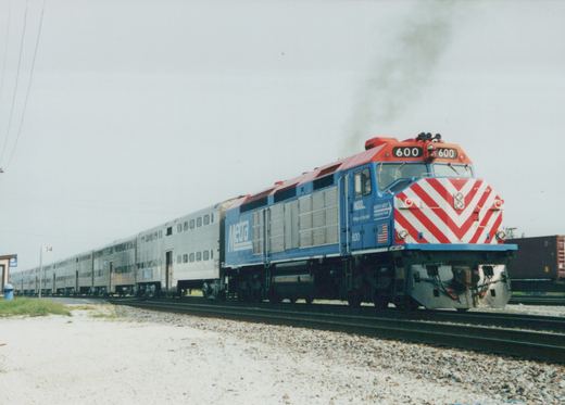 A blue and red Metra locomotive numbered 600 pulling a long line of silver passenger cars on a gravel track under an overcast sky.