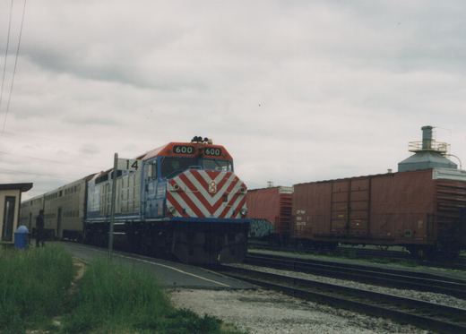A blue, gray, and orange Metra locomotive numbered 600 is at a station platform with commuter cars behind it, while a brown CP Rail boxcar sits on an adjacent track under an overcast sky.