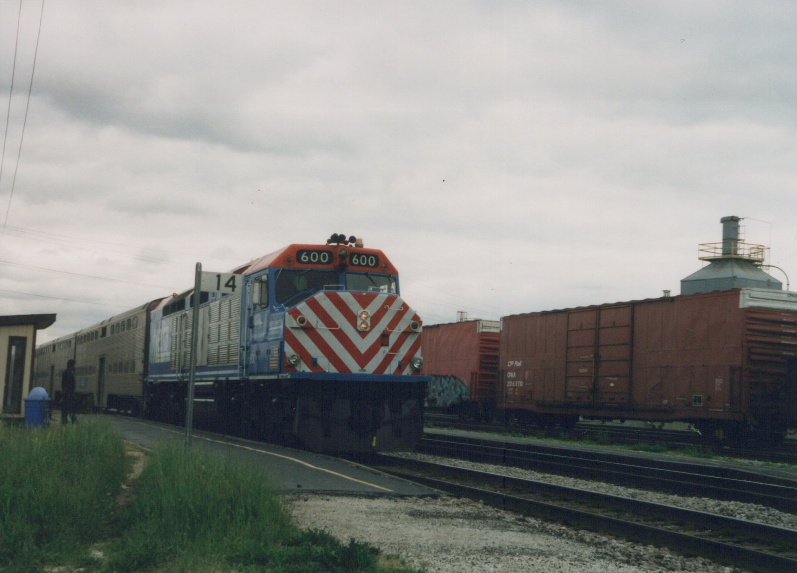 A blue, gray, and orange Metra locomotive numbered 600 is at a station platform with commuter cars behind it, while a brown CP Rail boxcar sits on an adjacent track under an overcast sky.