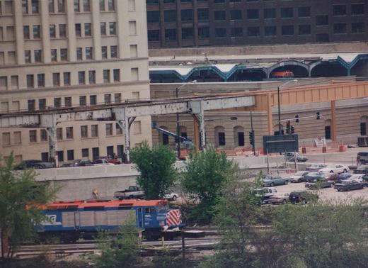 A blue and orange Metra locomotive, numbered 607, is on the tracks near a city street with cars and buildings in the background, under an overcast sky.