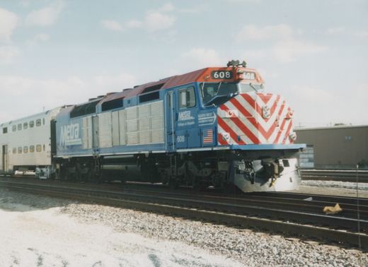 A blue and orange Metra diesel locomotive with the number 608 is coupled to a silver gallery car, sitting on tracks in a yard under a clear sky.