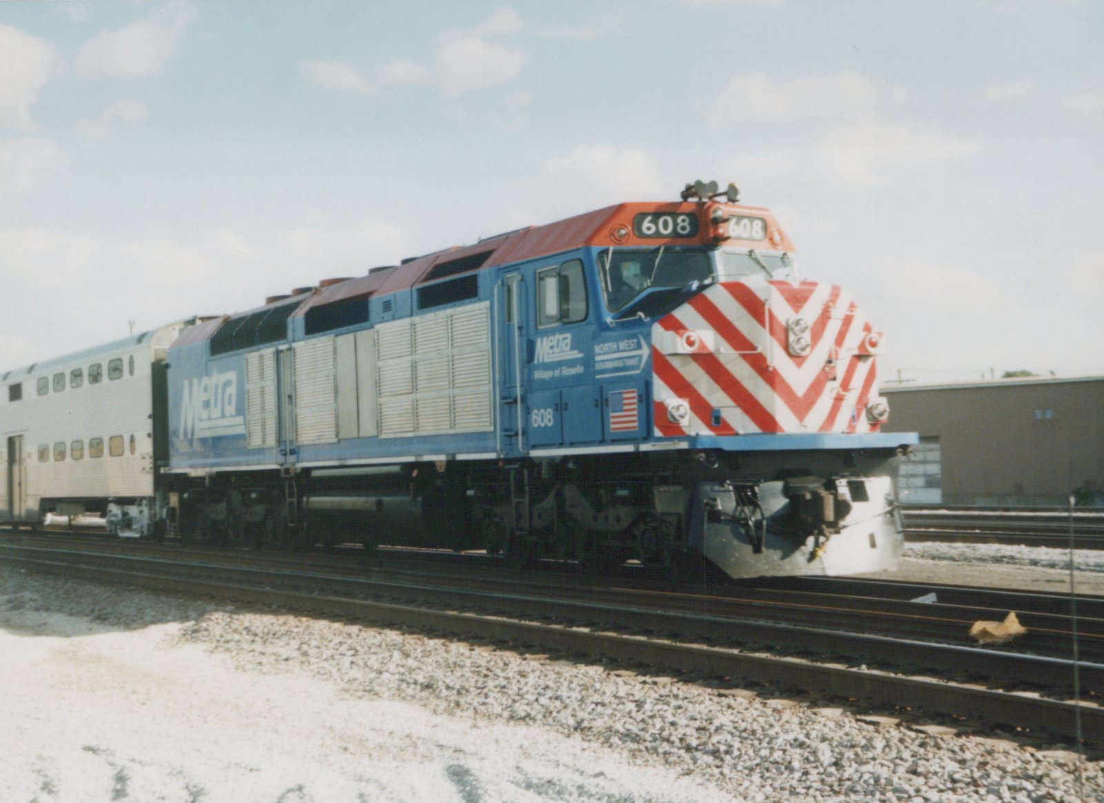 A blue and orange Metra diesel locomotive with the number 608 is coupled to a silver gallery car, sitting on tracks in a yard under a clear sky.