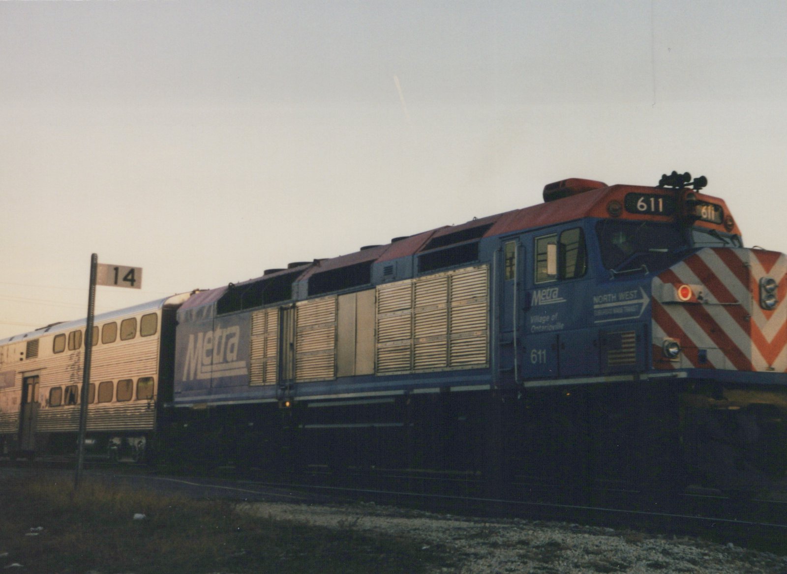 A blue and orange Metra locomotive numbered 611 is coupled to a silver double-decker passenger car, sitting on a track next to a grassy area under an overcast sky at dusk.