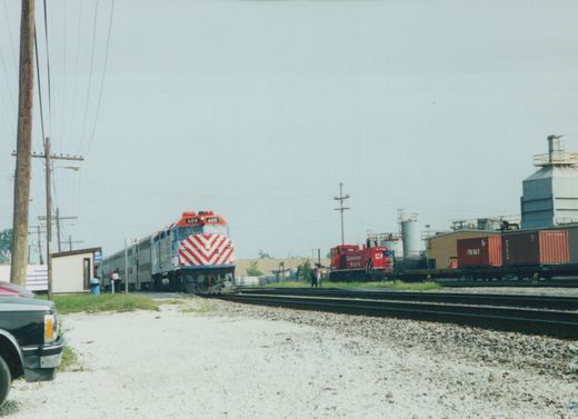 A red, white, and blue Metra locomotive #609 is parked on a set of tracks in a gravel yard, with a Canadian Pacific locomotive and some freight cars in the background under an overcast sky.