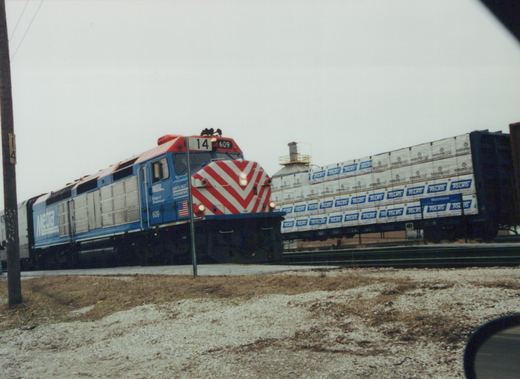 A blue and red Metra locomotive numbered 609 moves down a railroad track in winter, pulling freight cars with TOLKO branding, with snow on the ground and an overcast sky.