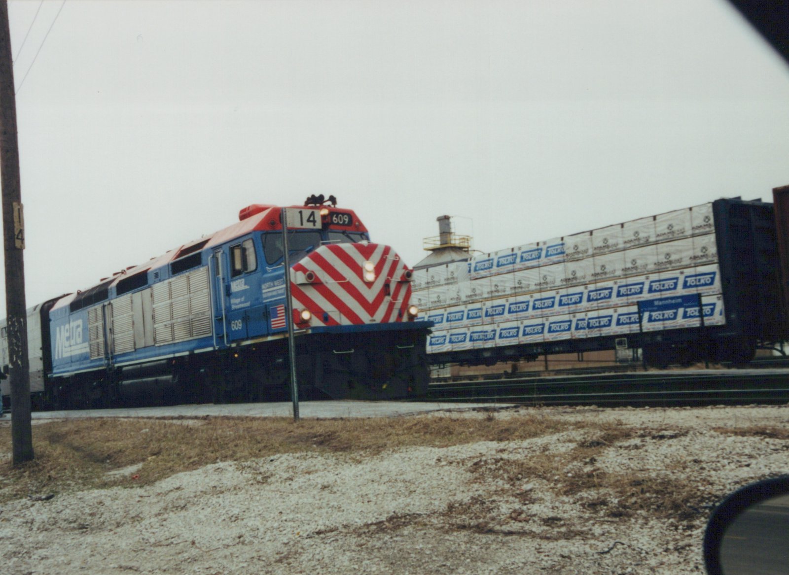 A blue and red Metra locomotive numbered 609 moves down a railroad track in winter, pulling freight cars with TOLKO branding, with snow on the ground and an overcast sky.