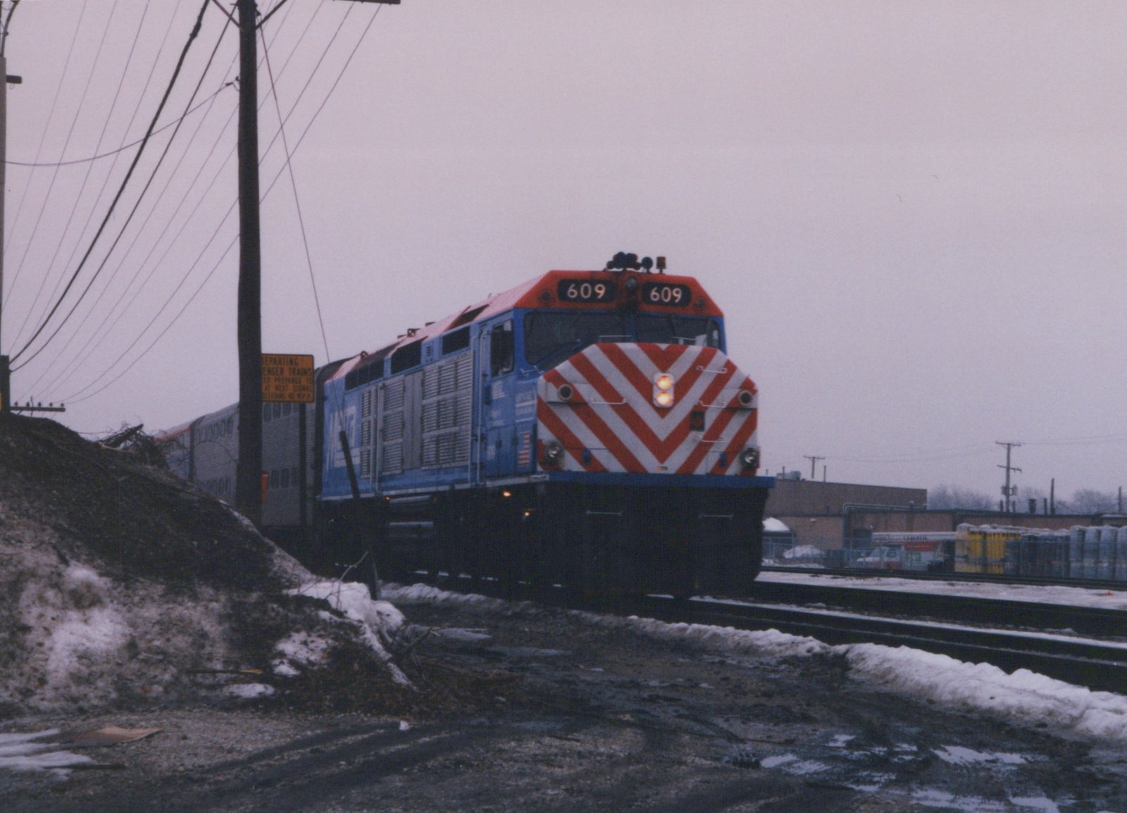 A blue Metra diesel locomotive numbered 609 leads a train of double-decker cars along a snow-dusted track, with an industrial backdrop and overcast sky.