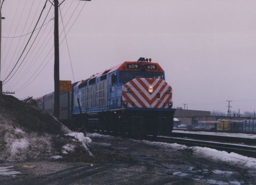 A blue Metra diesel locomotive numbered 609 leads a train of double-decker cars along a snow-dusted track, with an industrial backdrop and overcast sky.
