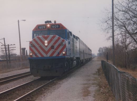 A Metra train with locomotive 609 and gallery cars travels along a private right-of-way, bordered by a chain-link fence and bare trees on a cloudy winter day.