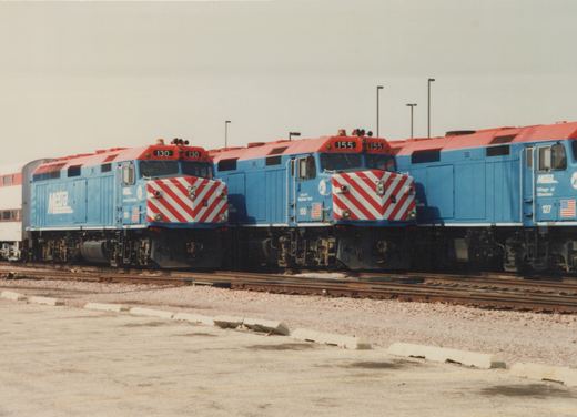 Three blue and red Metra locomotives with white chevrons on their noses are parked side by side on parallel tracks in a gravel yard under an overcast sky.