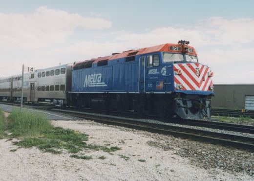 A blue Metra locomotive pulls silver double-decker commuter cars along railroad tracks beside a gravel and grassy area under a cloudy sky.