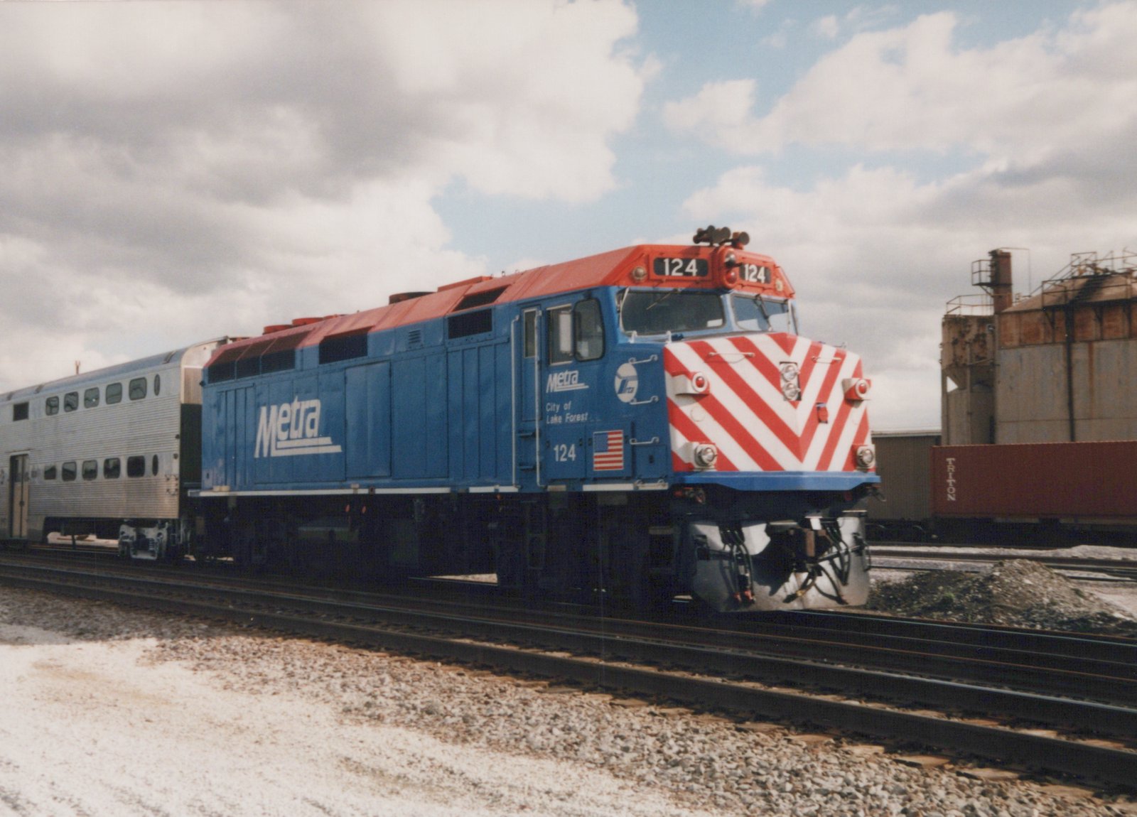 A blue and red Metra F40PH locomotive numbered 124 is shown pulling a silver double-decker commuter car on a snowy day, with industrial buildings in the background.