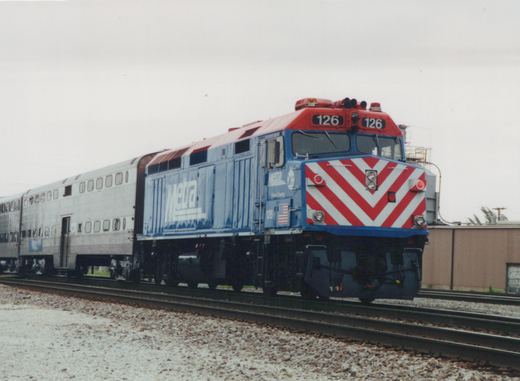 A blue Metra locomotive numbered 126 with a red and white chevron front is pulling silver passenger cars on a railroad track in a yard under an overcast sky.