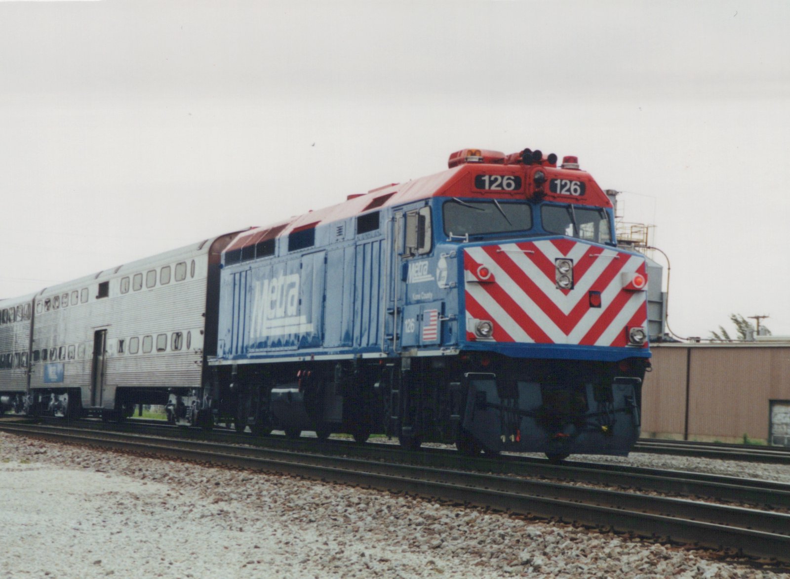 A blue Metra locomotive numbered 126 with a red and white chevron front is pulling silver passenger cars on a railroad track in a yard under an overcast sky.