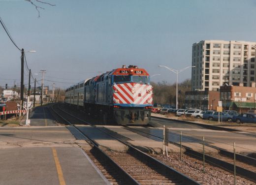 A Metra train with locomotive 126 and gallery cars crossing a street in a suburban area with a high-rise building in the background.