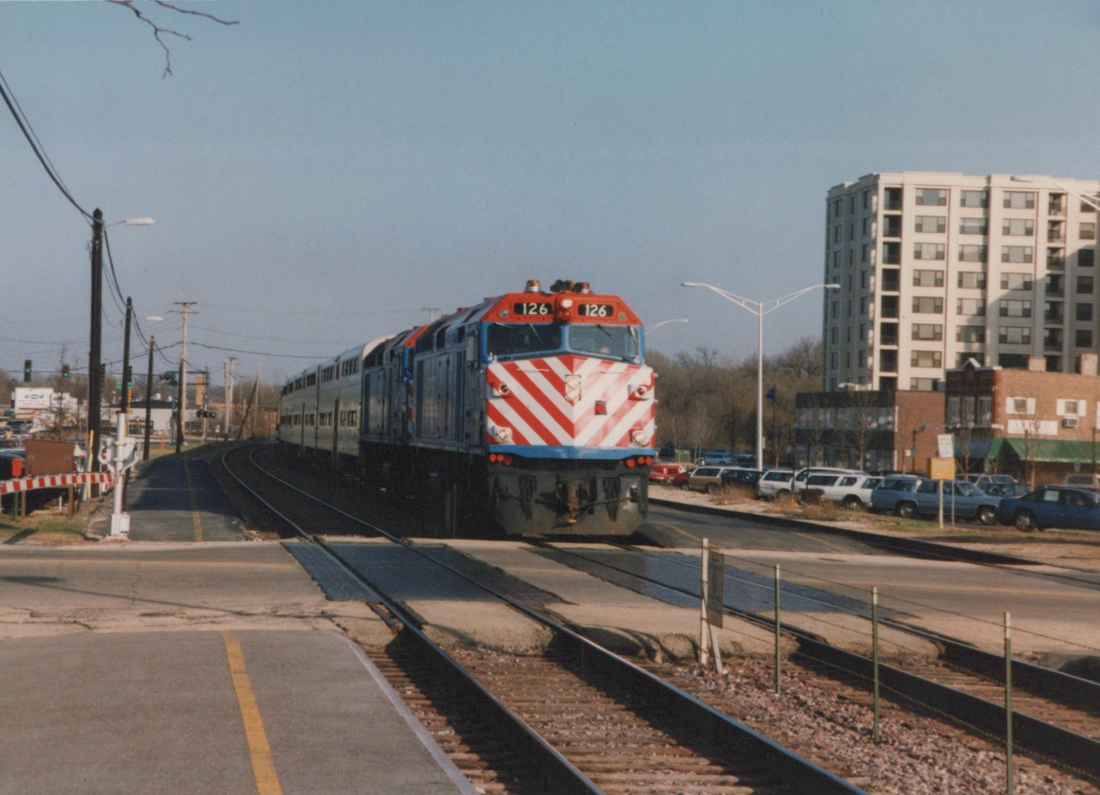 A Metra train with locomotive 126 and gallery cars crossing a street in a suburban area with a high-rise building in the background.