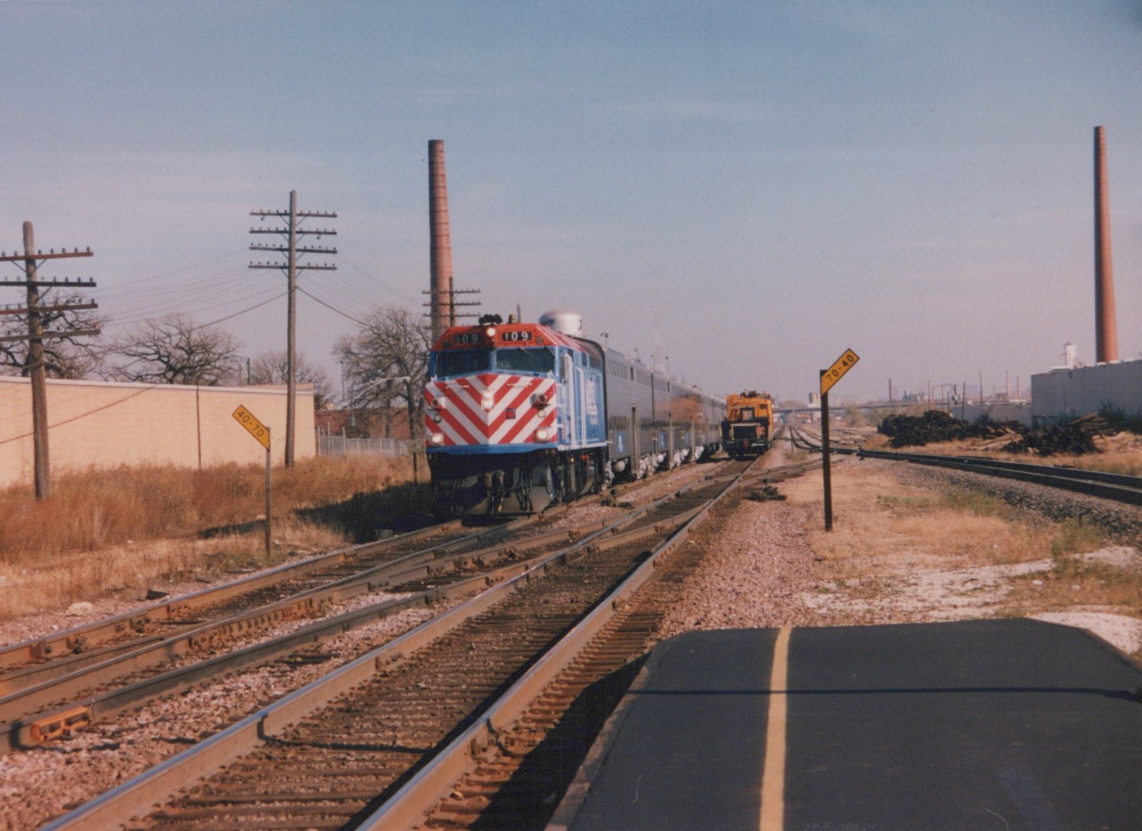 A blue and red Metra locomotive numbered 109 pulls a line of silver commuter cars along a set of tracks next to a road, with industrial buildings and smokestacks in the background under a clear sky.