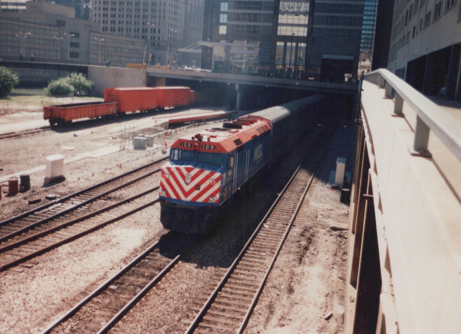 A blue and red Metra locomotive numbered 108 is shown from a high angle, traveling on tracks beneath a concrete overpass in a city setting with other freight cars nearby.