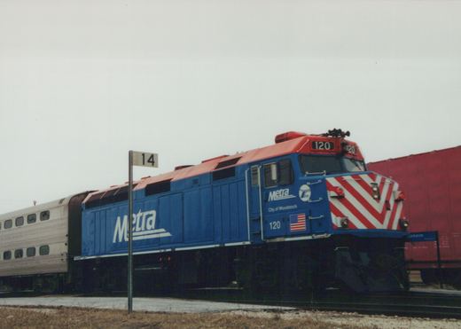 A blue and red Metra diesel locomotive with the number 120 and 'City of Woodstock' lettering on its side, pulling silver bi-level passenger cars at a grade crossing on an overcast, winter day.