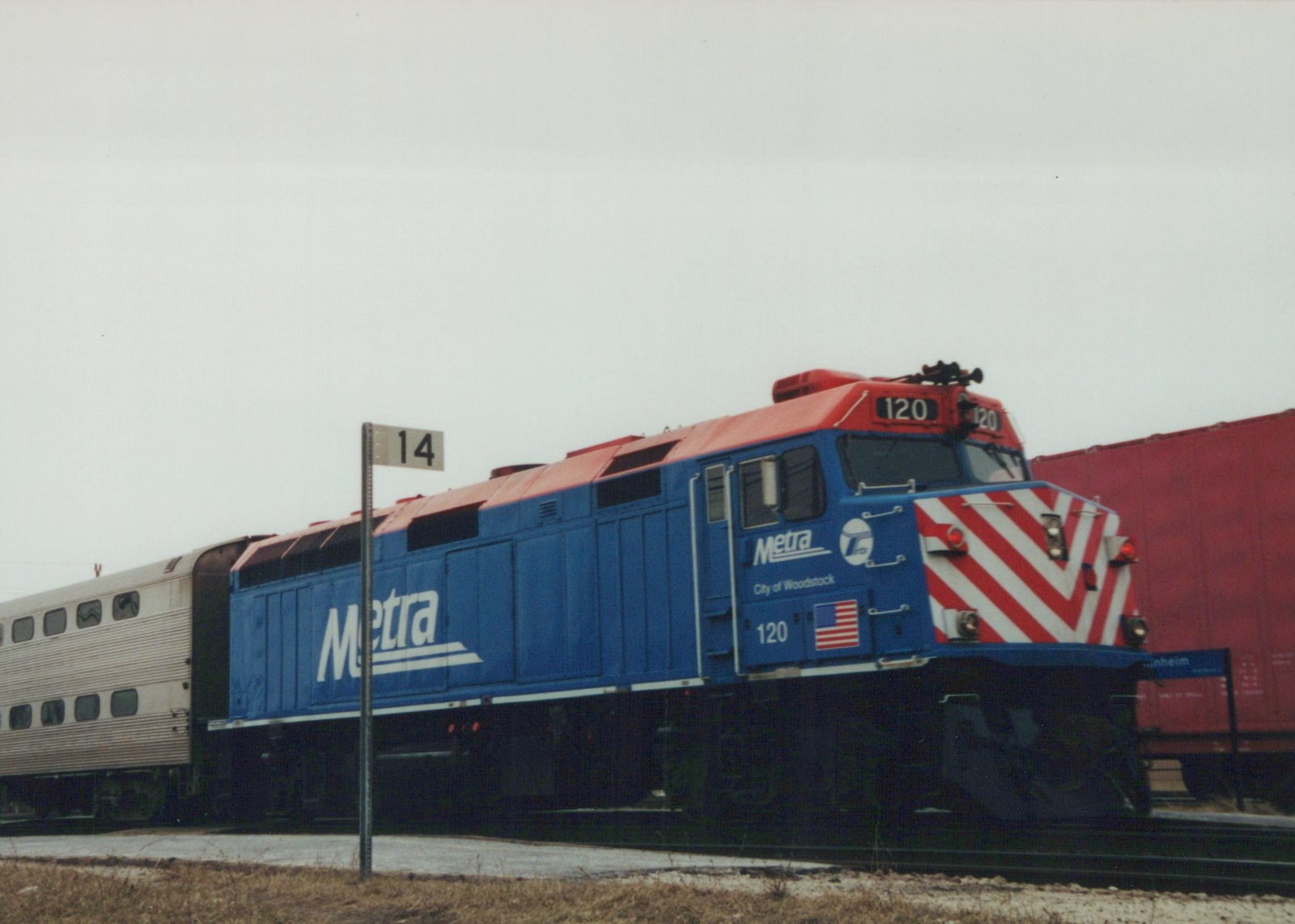 A blue and red Metra diesel locomotive with the number 120 and 'City of Woodstock' lettering on its side, pulling silver bi-level passenger cars at a grade crossing on an overcast, winter day.