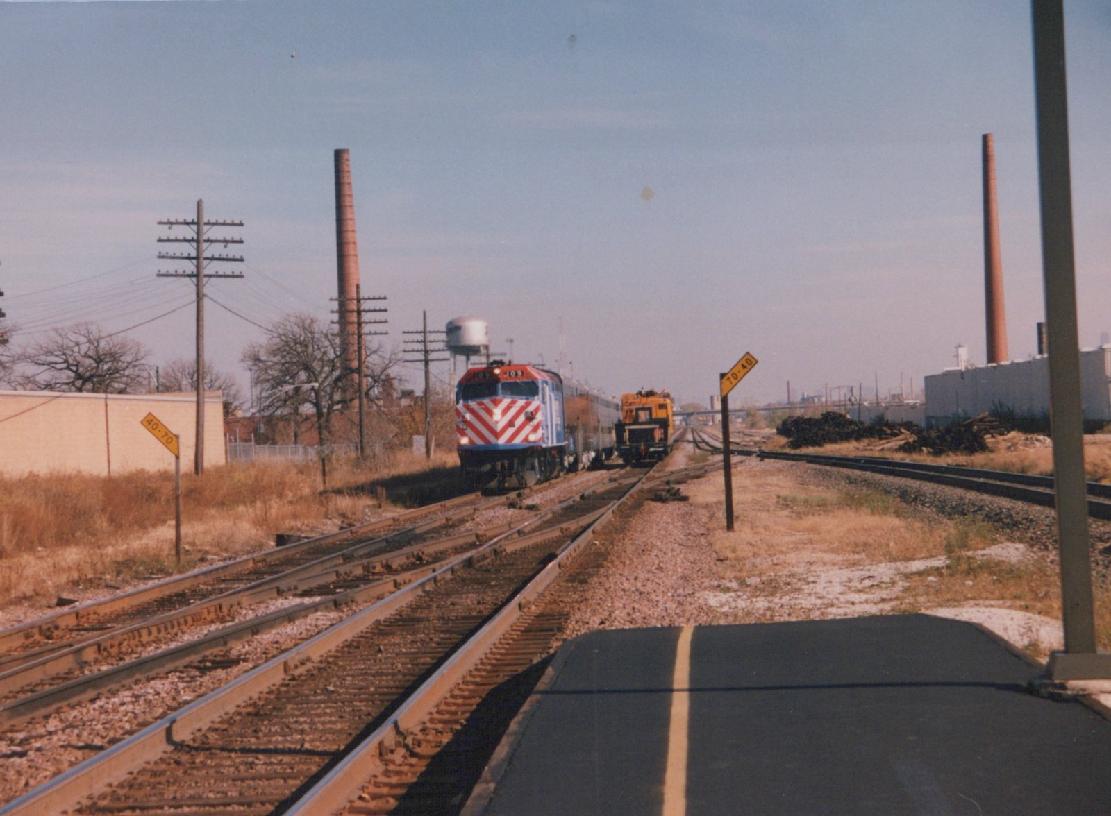 A Metra train with a red, white, and blue locomotive numbered 409 travels on tracks beside a road and industrial buildings under a clear autumn sky.