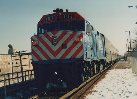 A blue, red, and white Metra diesel locomotive numbered 107 is at a station platform with snow on the ground, pulling a train of stainless steel passenger cars.