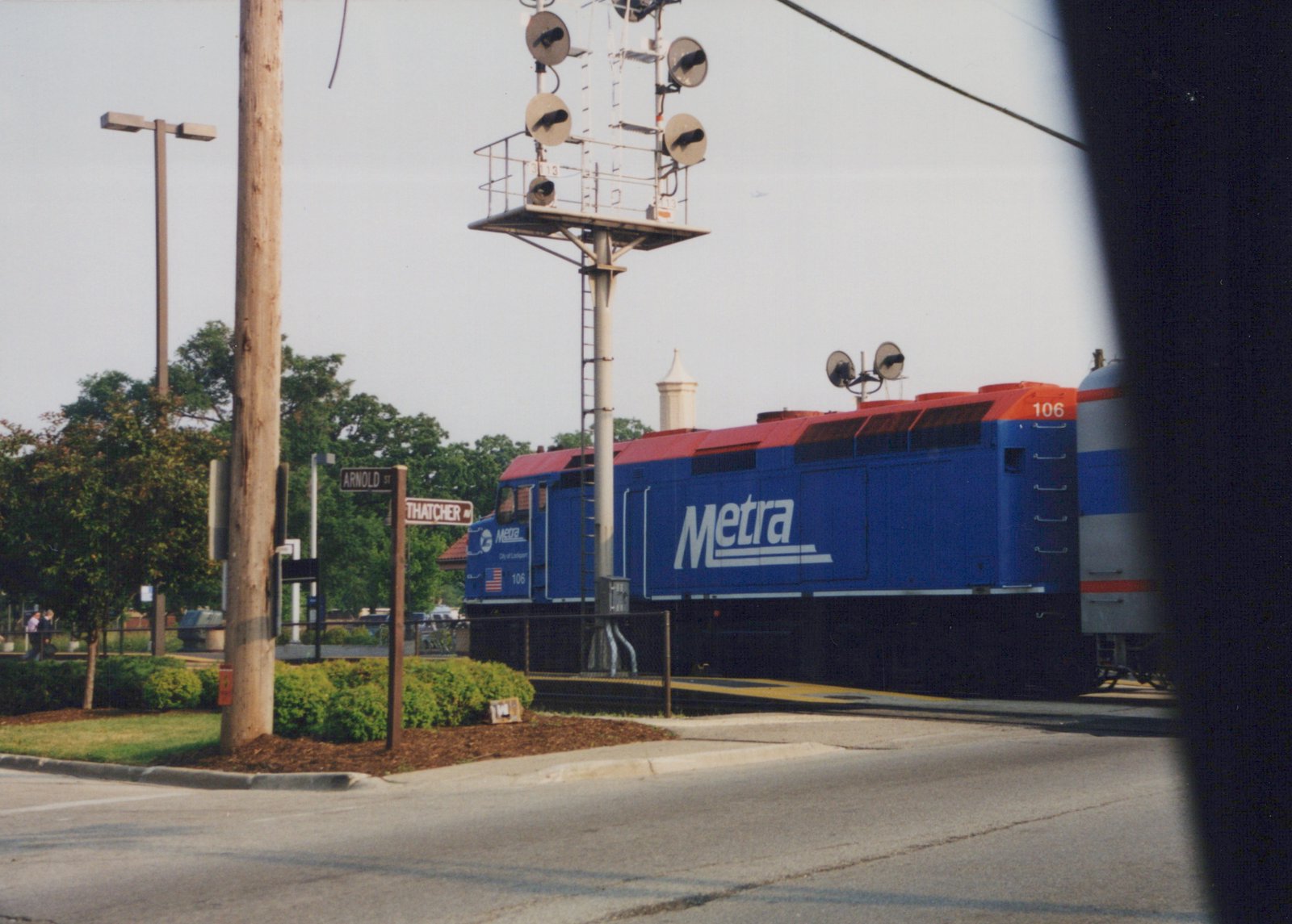 A blue and red Metra diesel locomotive numbered 106 sits at a station platform next to street signs for Arnold and Thatcher streets under a clear sky.