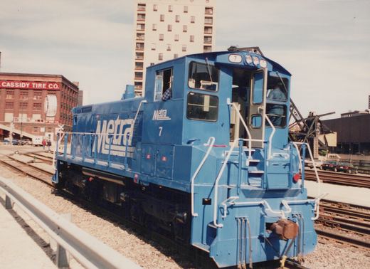 A bright blue Metra locomotive, number 7, is stopped on railroad tracks near a street with a brick building and a tall apartment building in the background.