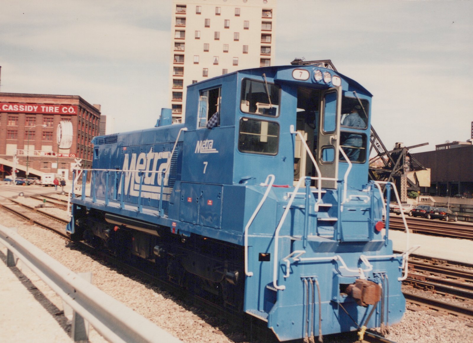 A bright blue Metra locomotive, number 7, is stopped on railroad tracks near a street with a brick building and a tall apartment building in the background.