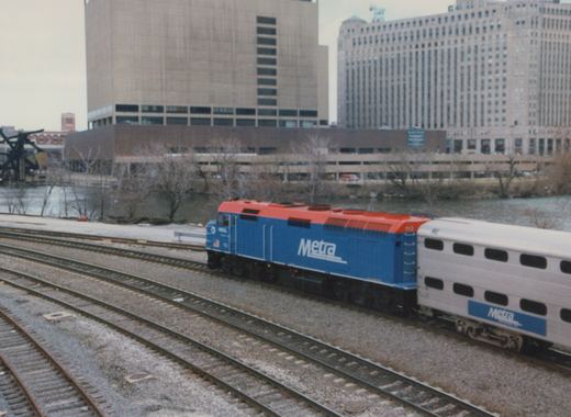 A blue and orange Metra locomotive numbered 112 pulls a silver double-decker passenger car on multiple train tracks next to a river; bare trees and snow are visible, with tall buildings in the background.