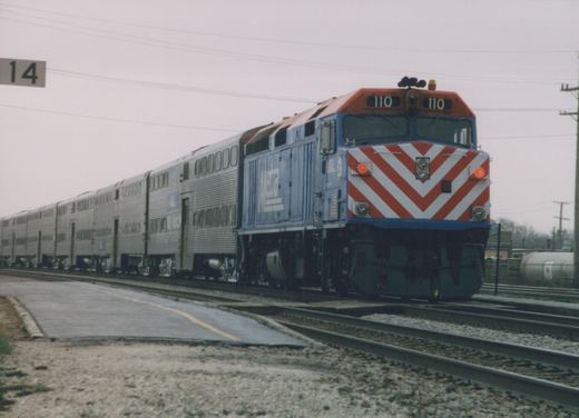 A blue and orange Metra diesel locomotive with the number 110 on its front, pulling a line of silver passenger cars on a set of tracks under an overcast sky.