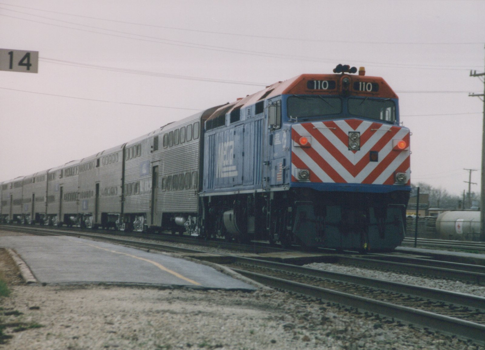A blue and orange Metra diesel locomotive with the number 110 on its front, pulling a line of silver passenger cars on a set of tracks under an overcast sky.