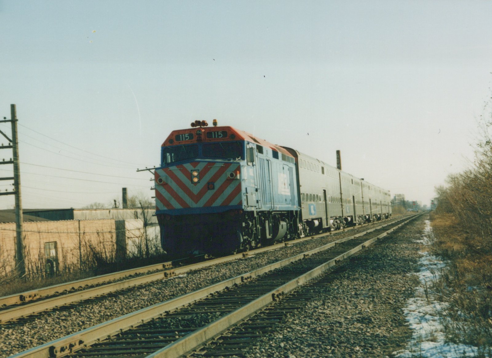 A Metra train with locomotive number 115, painted in blue with red and white stripes on the front, travels along a set of tracks next to a road with some snow on the ground and bare trees.