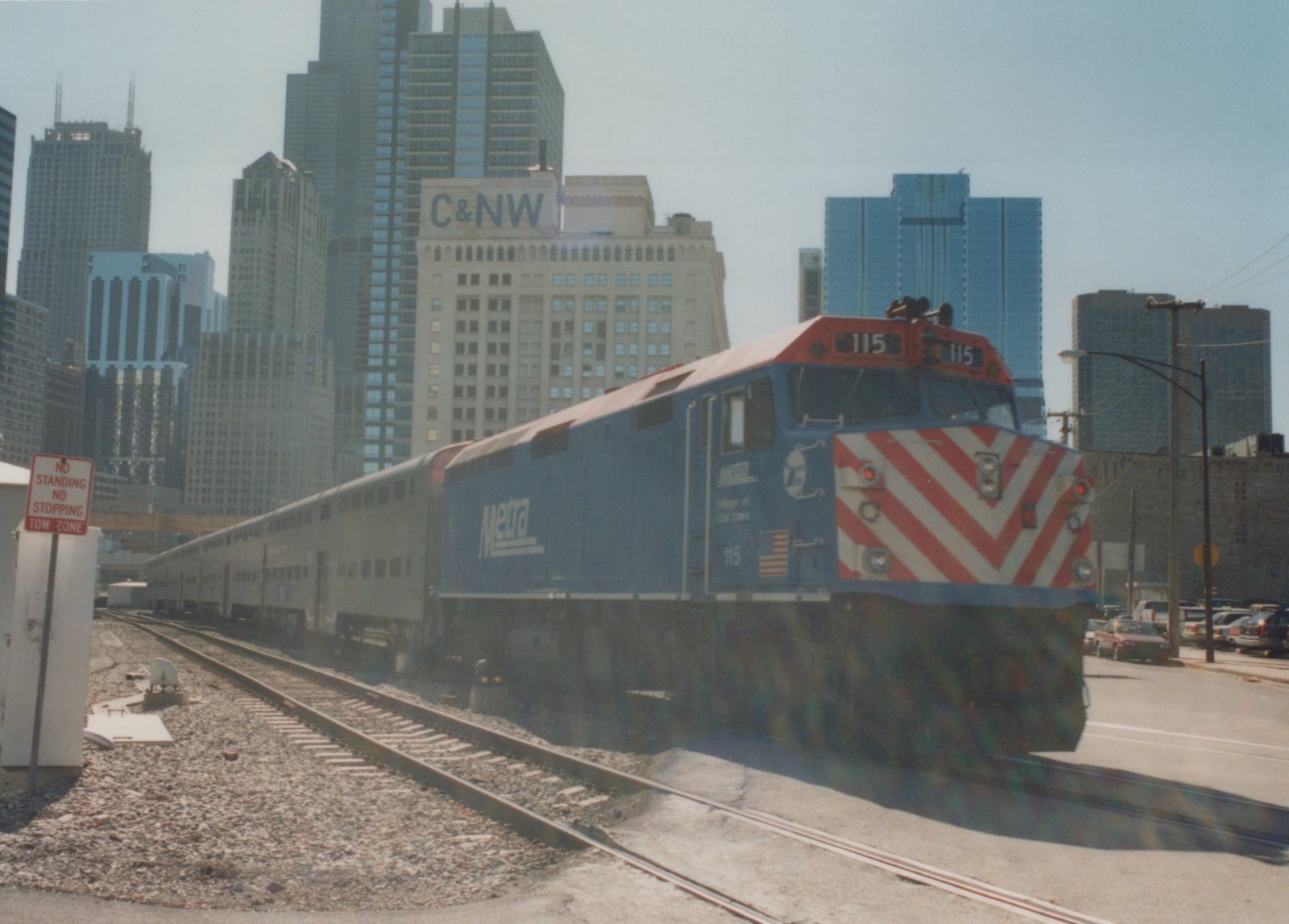 A blue Metra diesel locomotive numbered 115 pulls a line of silver passenger cars on a sunny day in downtown Chicago, with tall buildings and a street sign visible.